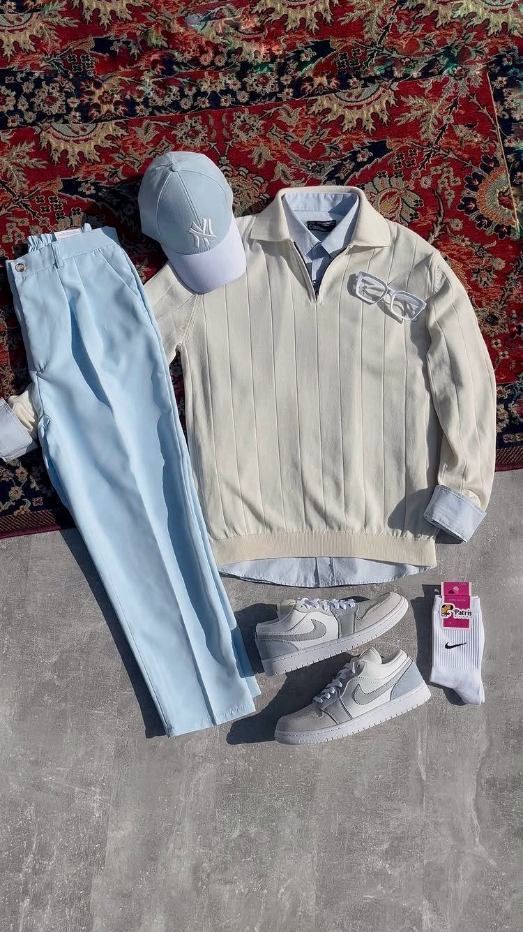 Light blue trousers, a cream striped sweater, a blue cap, and white sneakers laid out on a rug.