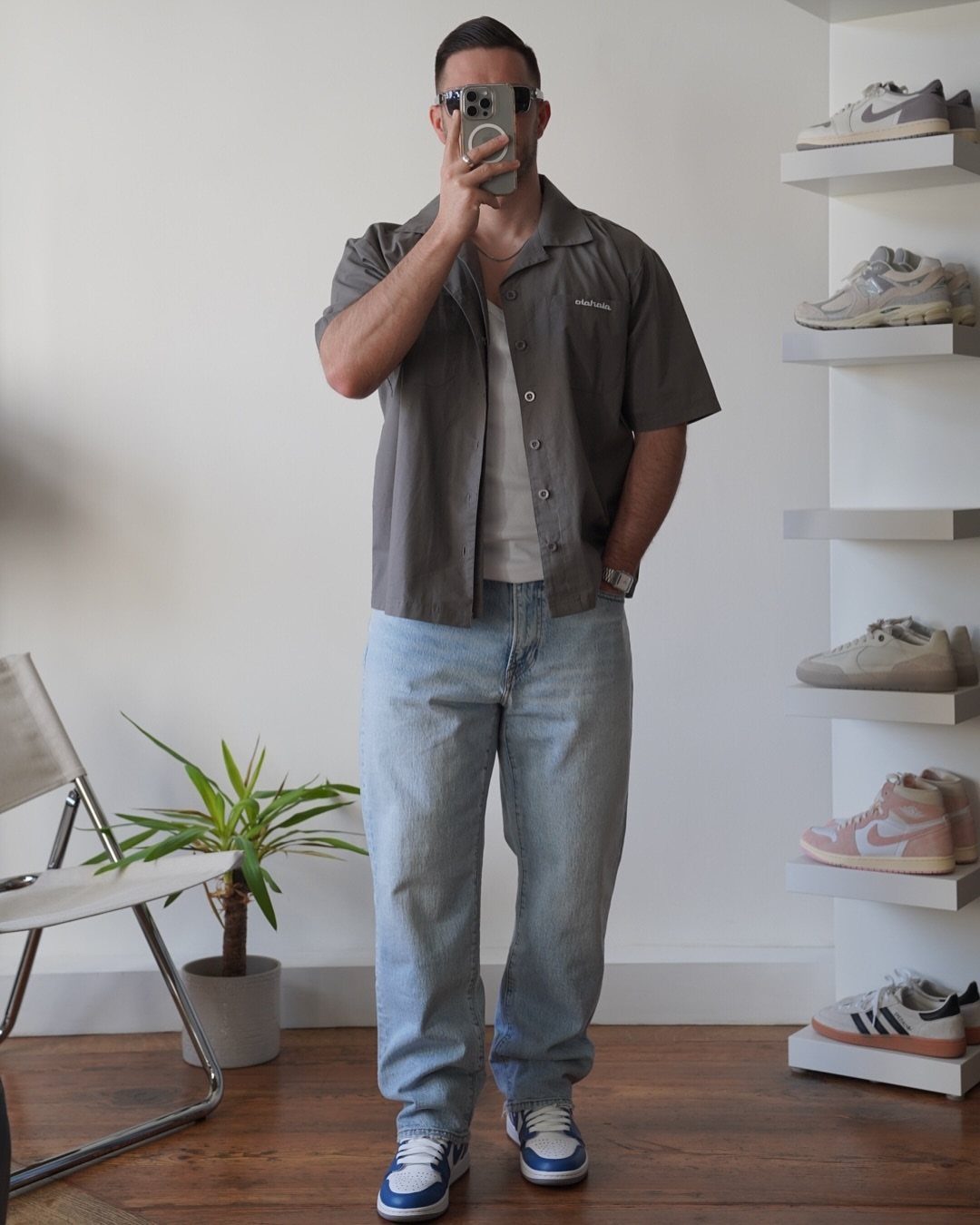 A man posing in a grey shirt, white tank top, light blue jeans, and blue sneakers.