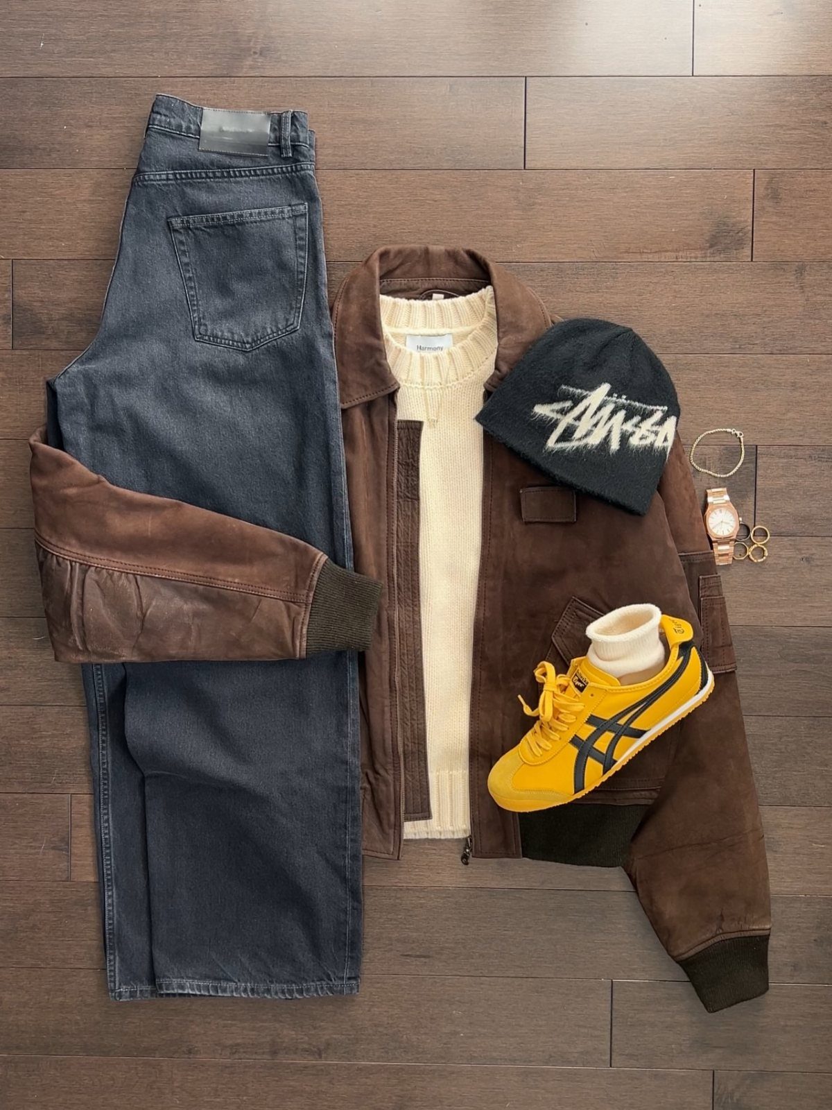 Men's spring outfit flat lay featuring blue jeans, brown jacket, cream sweater, black beanie, and yellow sneakers.