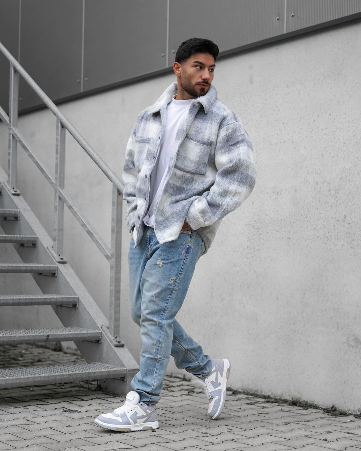 Man in a patterned flannel shirt, light blue jeans, and grey sneakers walking outdoors.