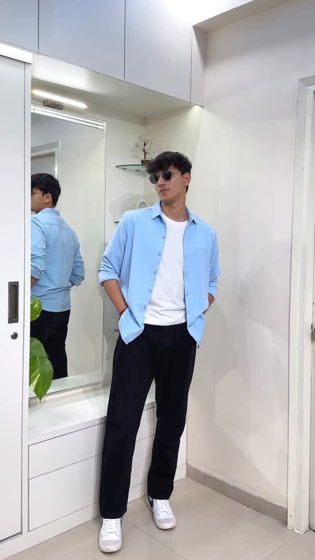 Man wearing a light blue open shirt over a white t-shirt with black pants.