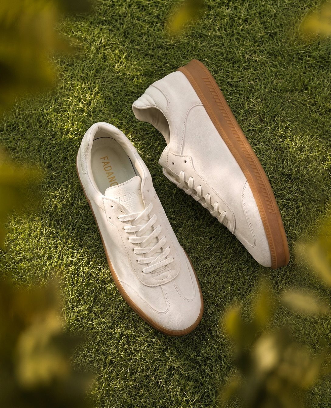 Cream suede minimalist sneakers with tan gum soles resting on green grass.