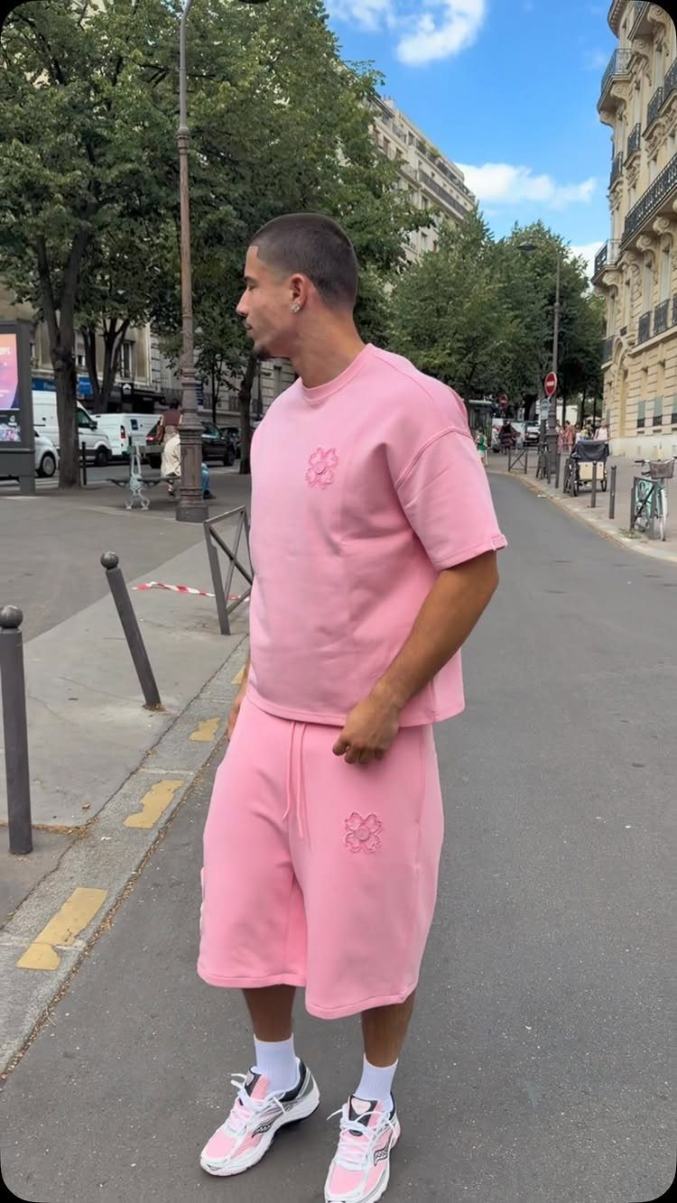 A man wearing a matching pink oversized t-shirt and shorts set with white sneakers standing on a city street.