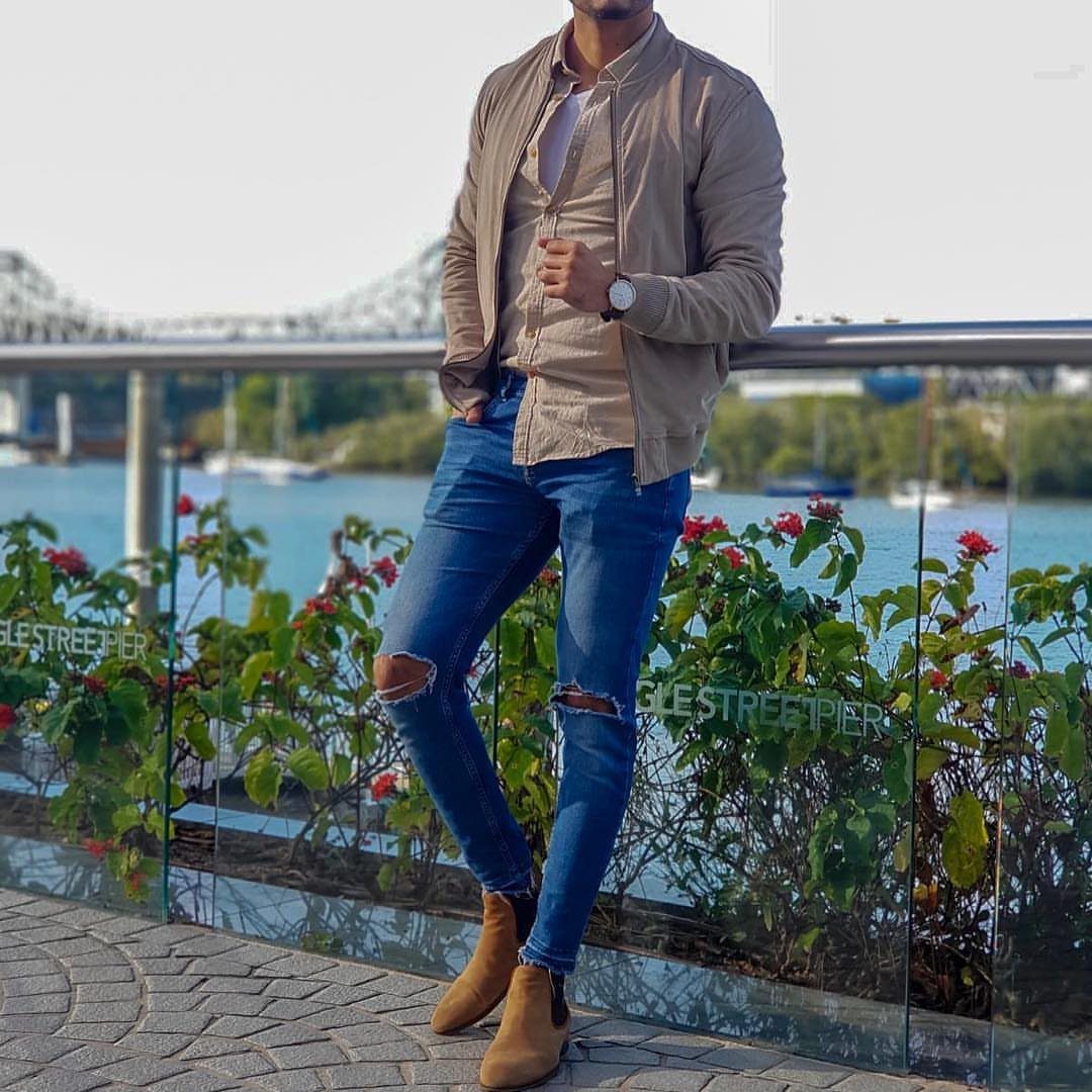 Man in tan suede bomber jacket, ripped blue jeans, and tan suede boots leaning on a railing.
