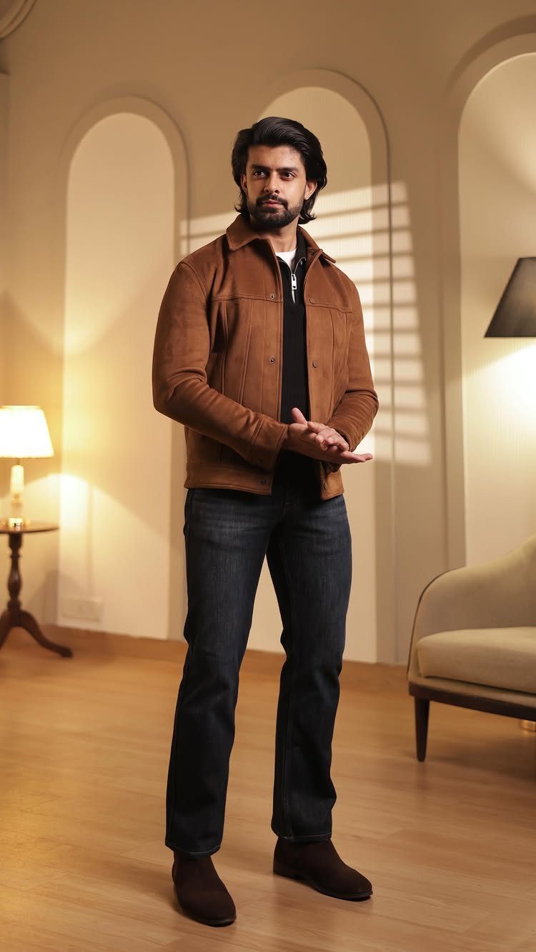 Man wearing a brown suede jacket, dark jeans, and brown suede ankle boots standing in a room.