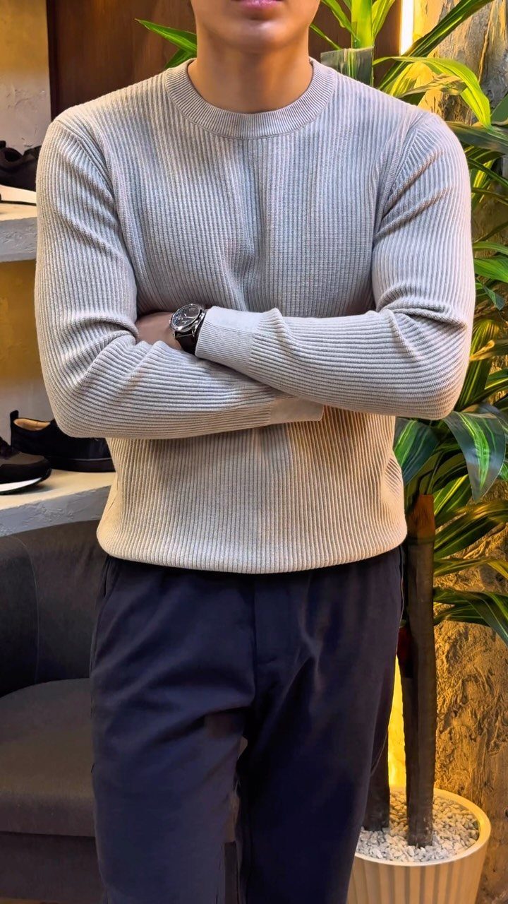 A man wearing a beige ribbed crew neck sweater and navy blue chinos standing with arms crossed.