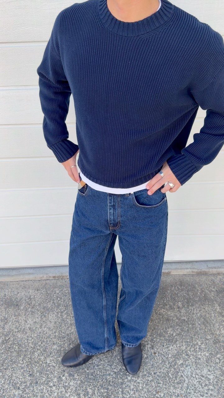Man wearing a navy blue ribbed sweater, baggy blue jeans, and black boots standing against a white wall.