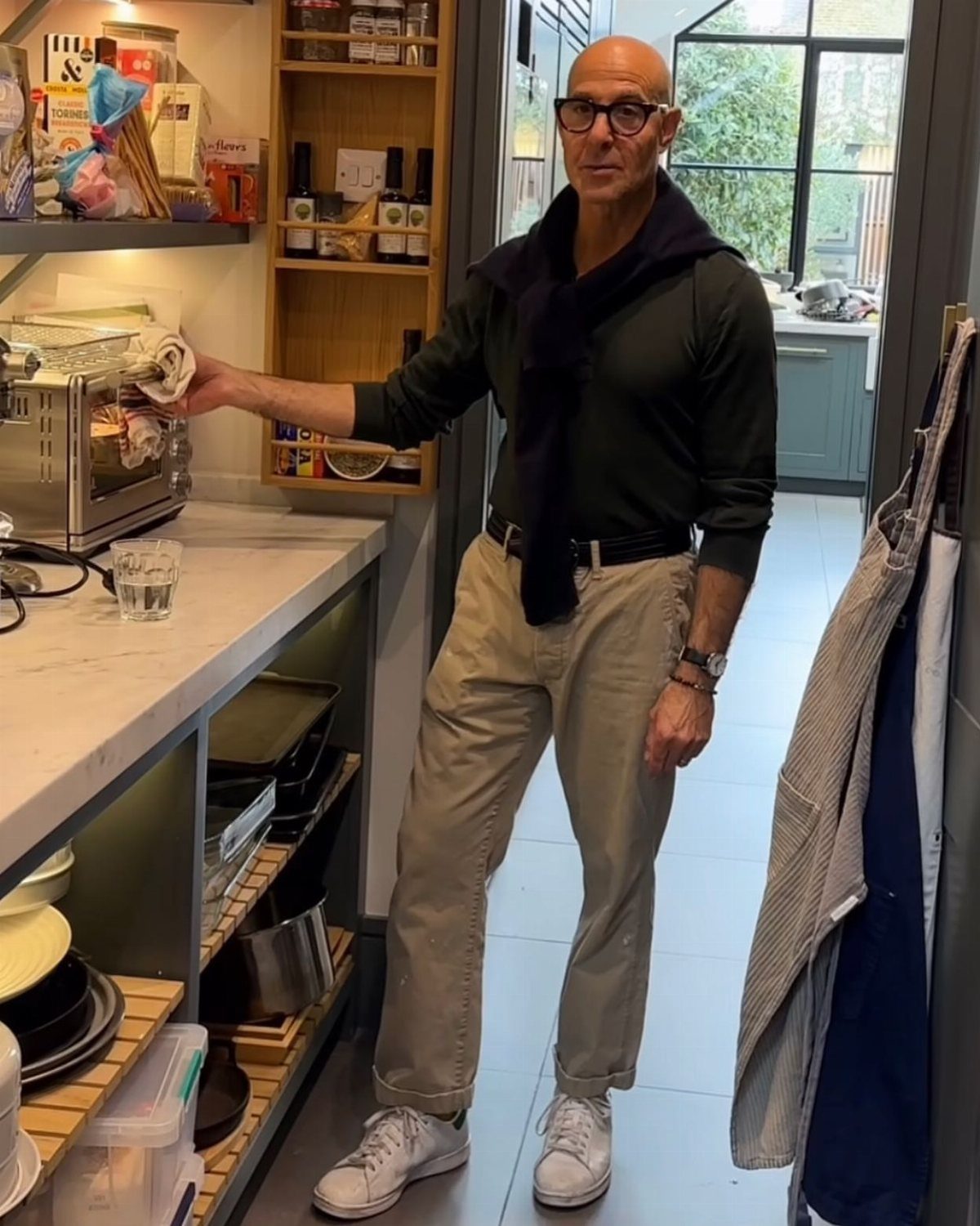 A man in a dark green sweater and beige chinos standing in a kitchen.