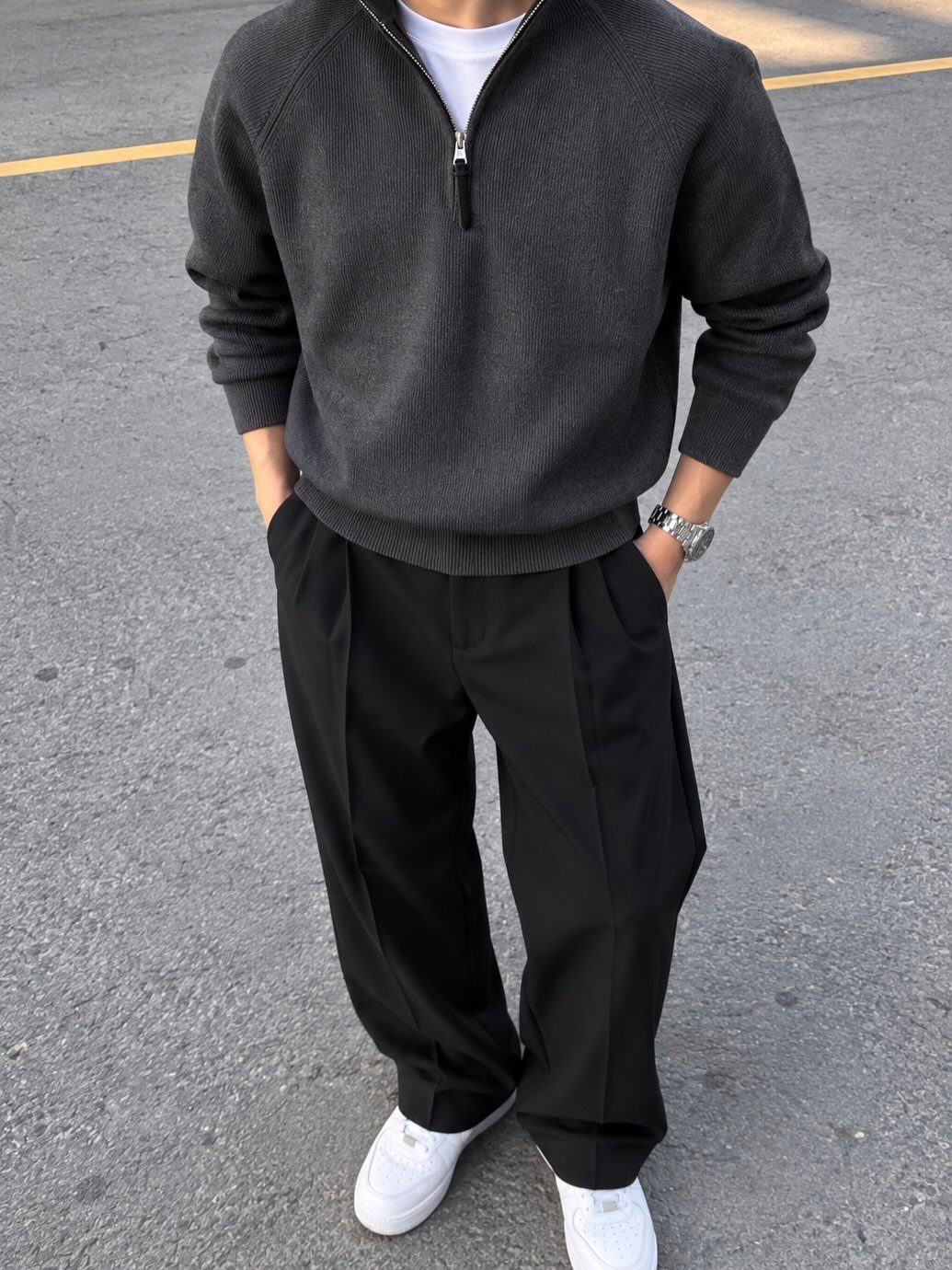 Man wearing a charcoal grey quarter-zip sweater, black pleated trousers, and white sneakers standing on asphalt.
