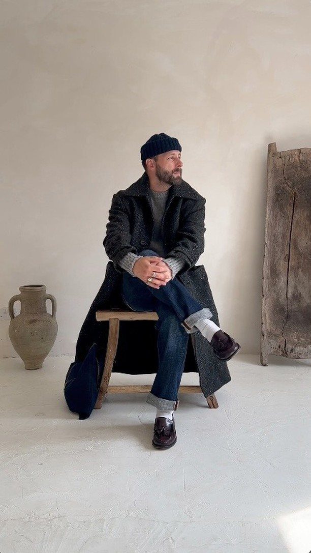 Man in a dark crew neck sweater, long coat, and blue jeans sitting on a wooden stool.