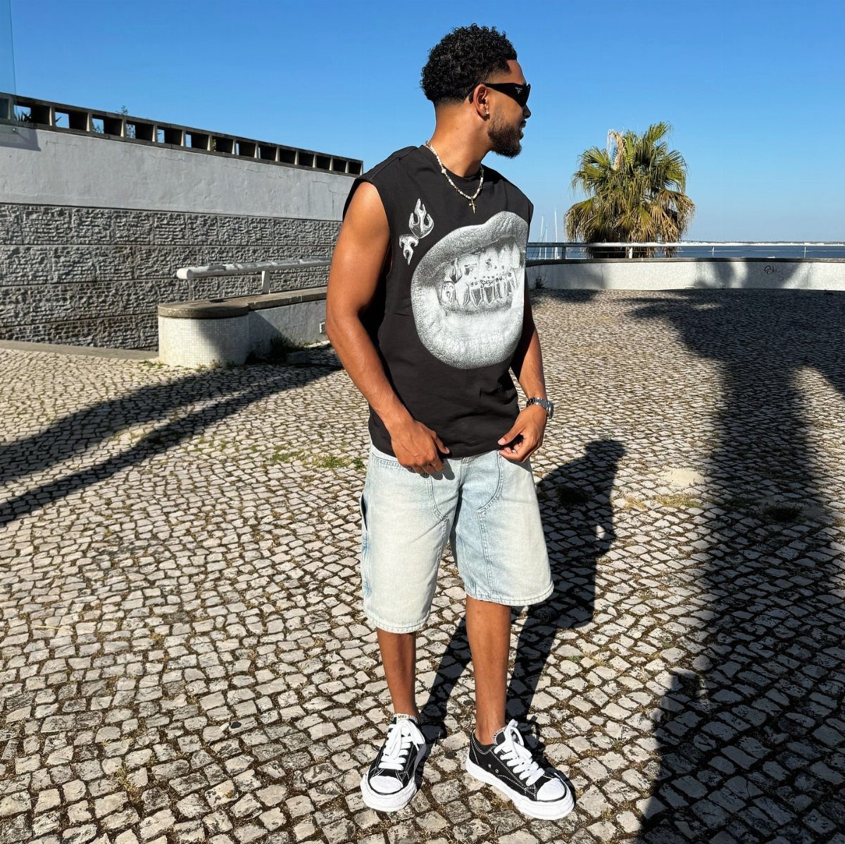A man posing in a black graphic tank top, denim shorts, and sneakers on a cobblestone ground.