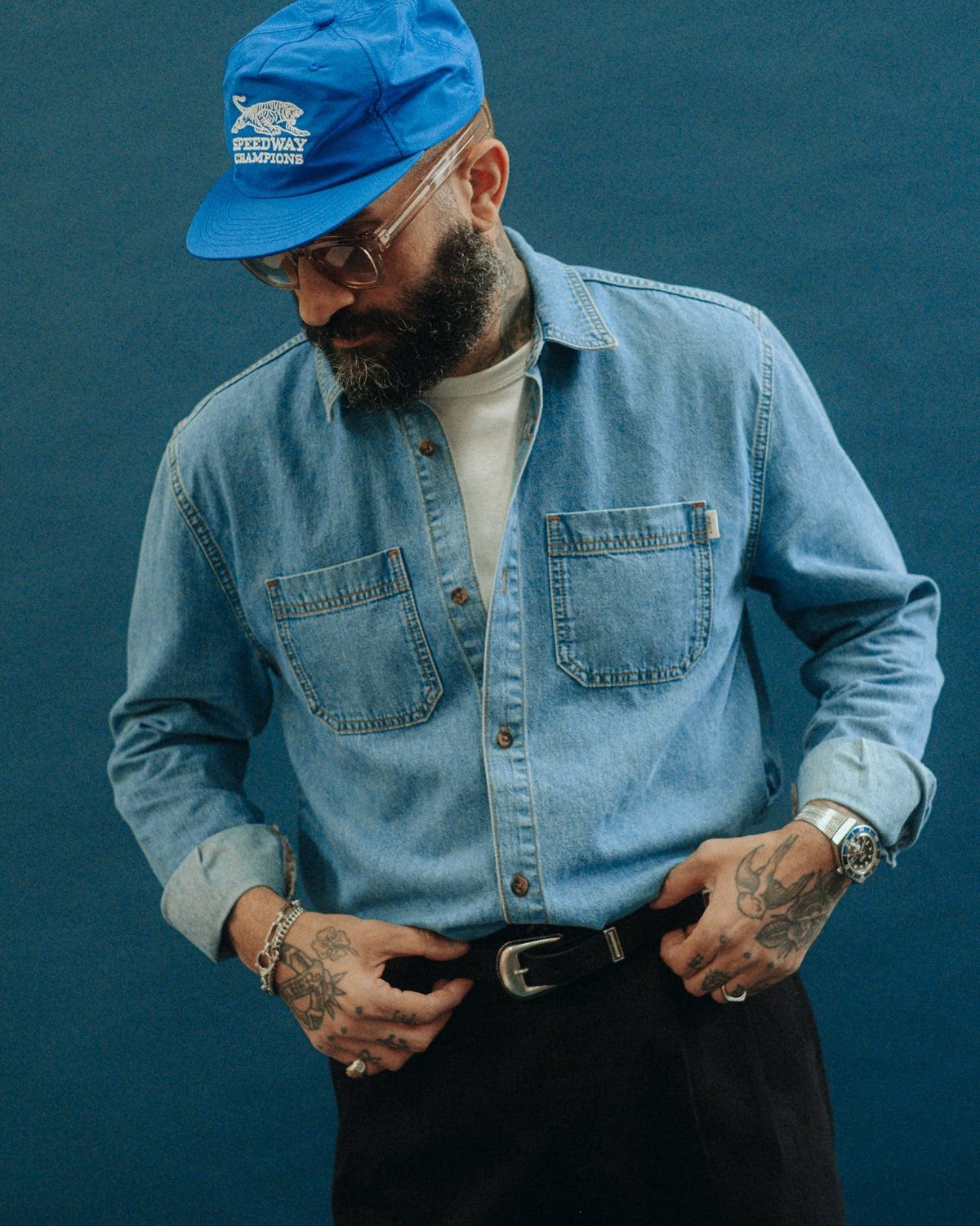 A bearded man wearing a white tank top under an unbuttoned denim shirt.