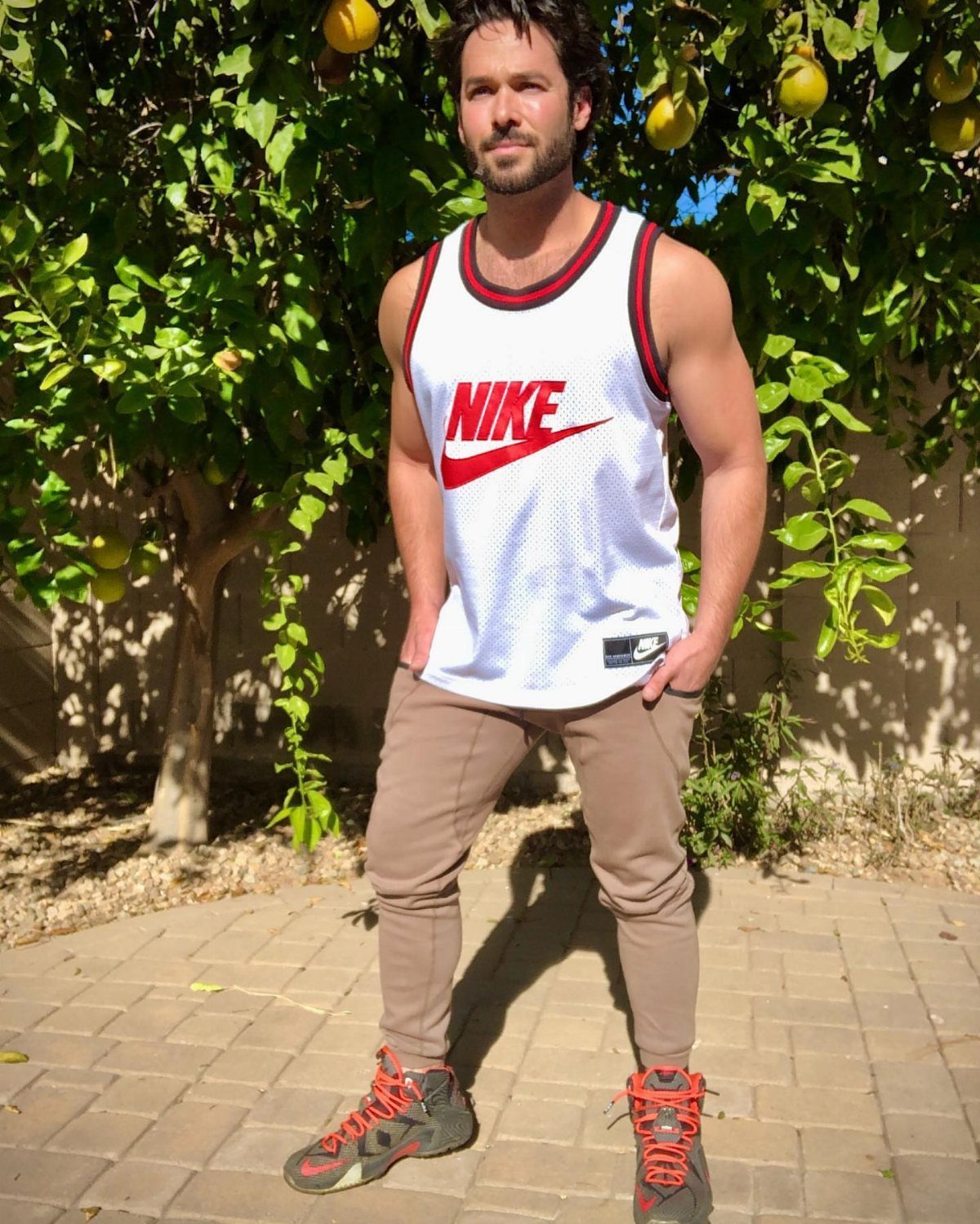 Man wearing a white Nike tank top, tan joggers, and red sneakers standing by a tree.