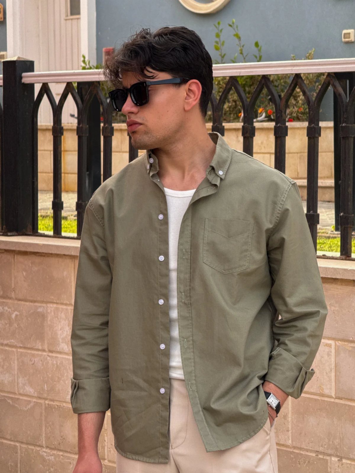 A man wearing an open olive shirt over a white ribbed tank top and beige pants.