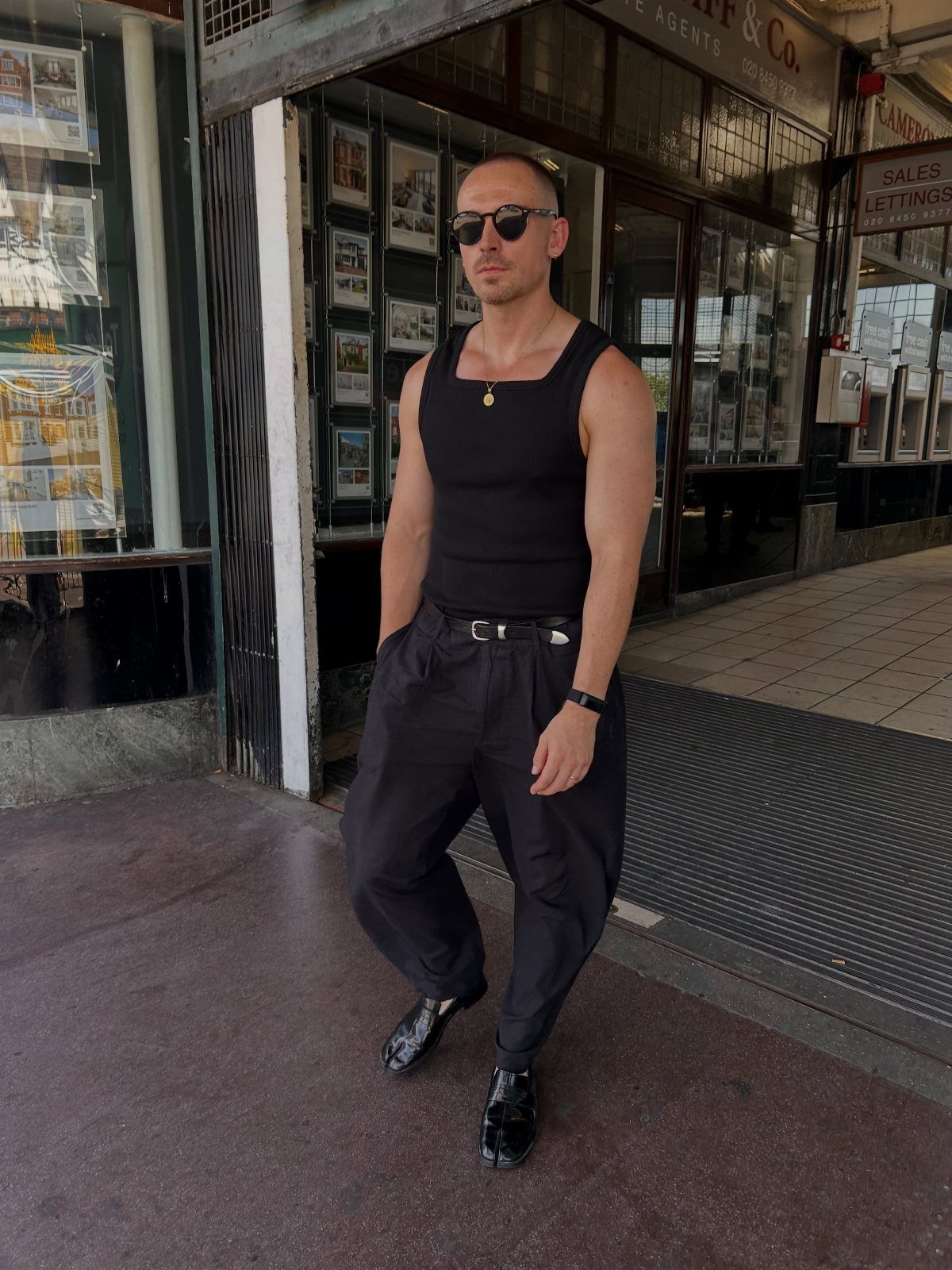 A man posing in a black tank top and black tailored trousers with sunglasses.