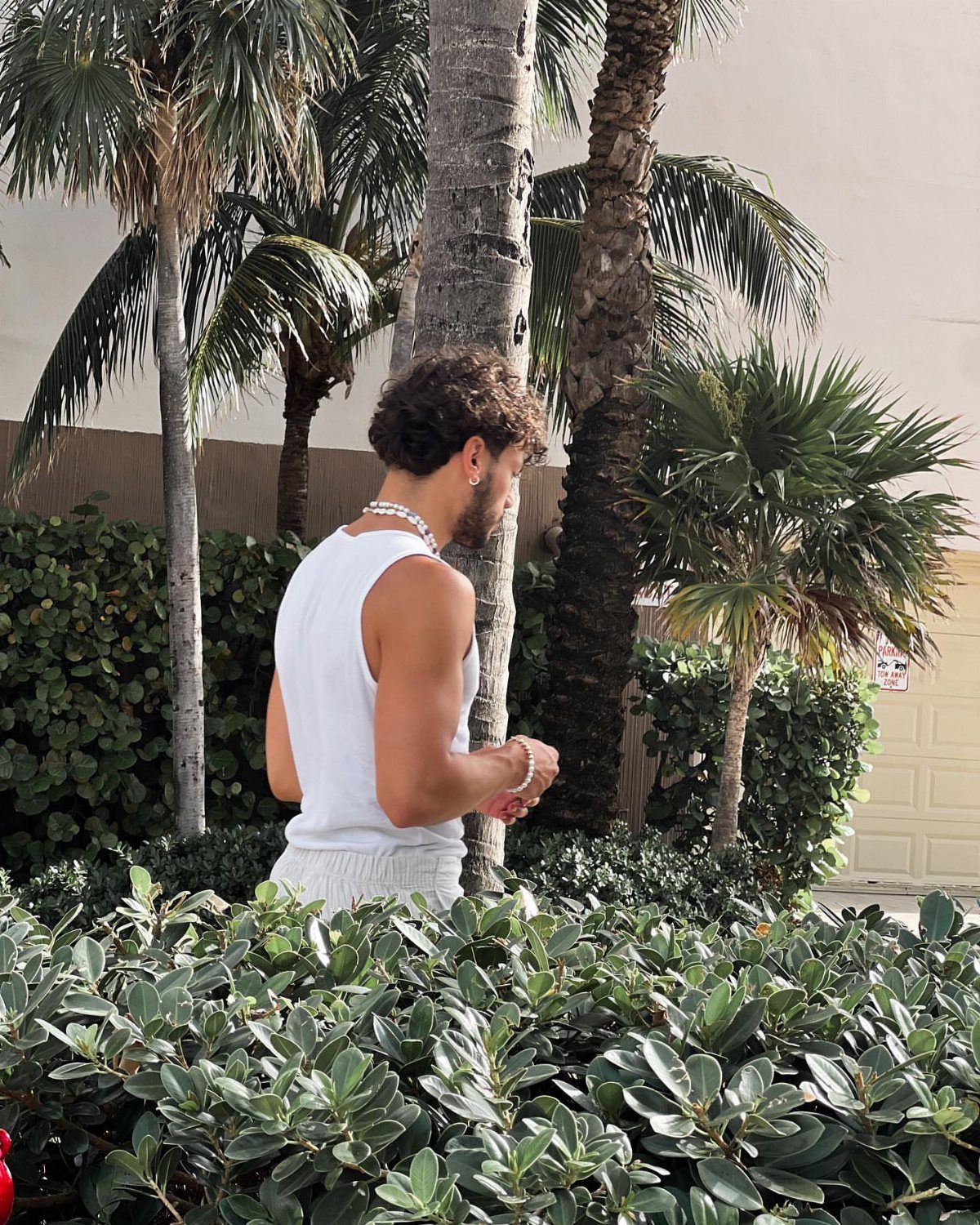 A man in a white tank top and shorts standing among palm trees and lush greenery.