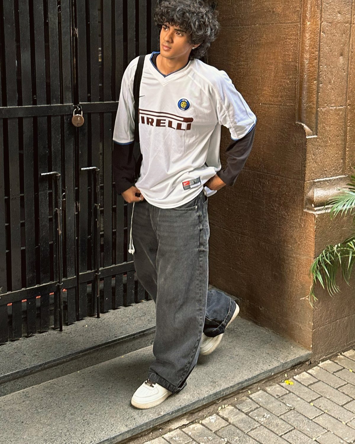 Man posing in a white Pirelli soccer jersey, black long-sleeve shirt, and baggy grey jeans.
