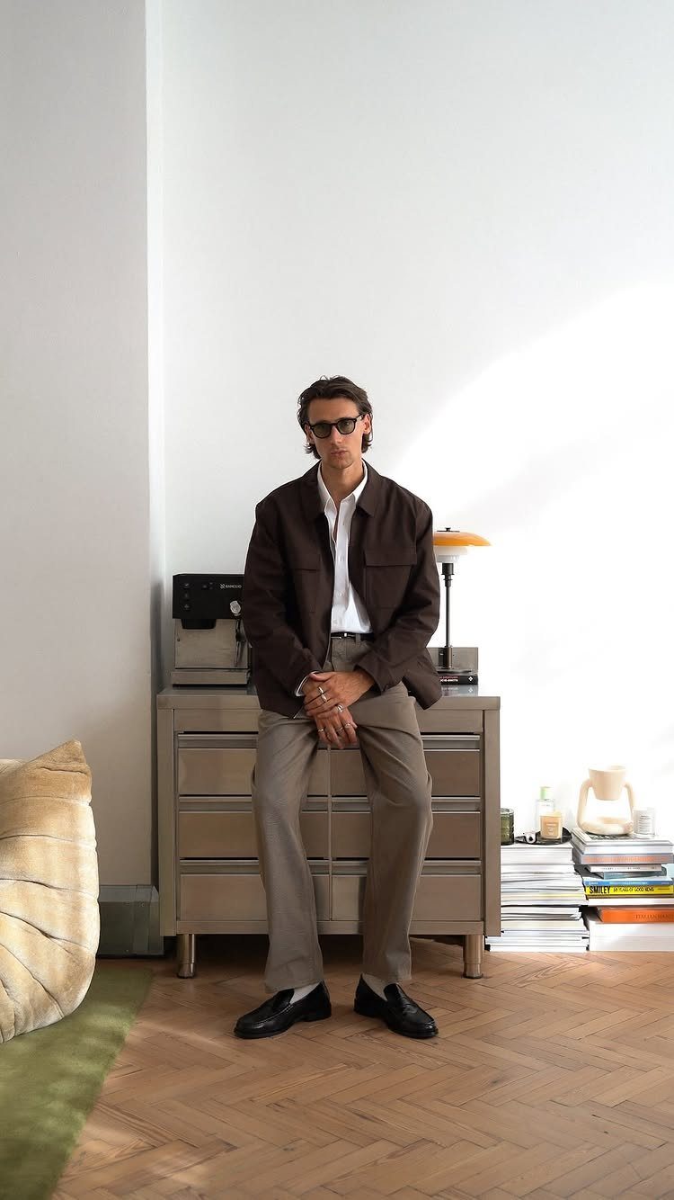 A man in a brown jacket, white shirt, and tan trousers sitting on a dresser.