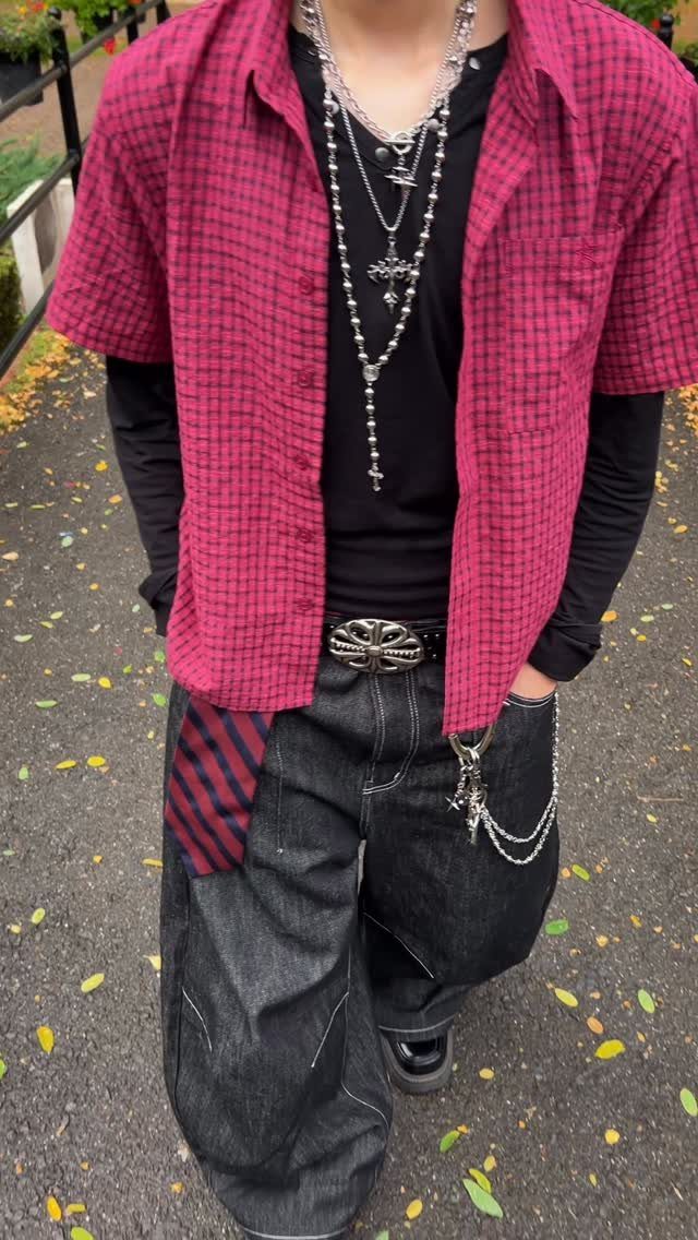 A person wearing a pink checkered shirt, black long-sleeve shirt, baggy black jeans, and silver jewelry.