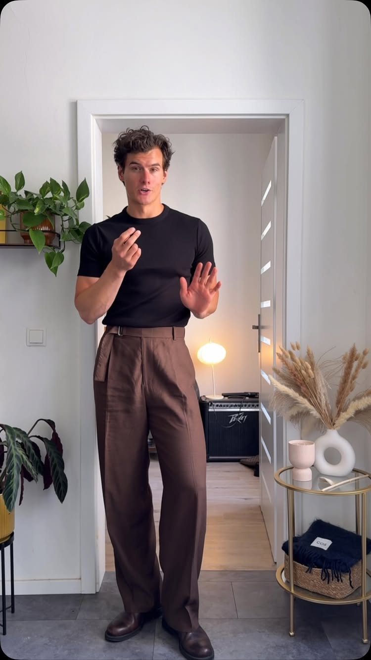 Man wearing a black t-shirt and brown wide-leg trousers standing in a doorway.