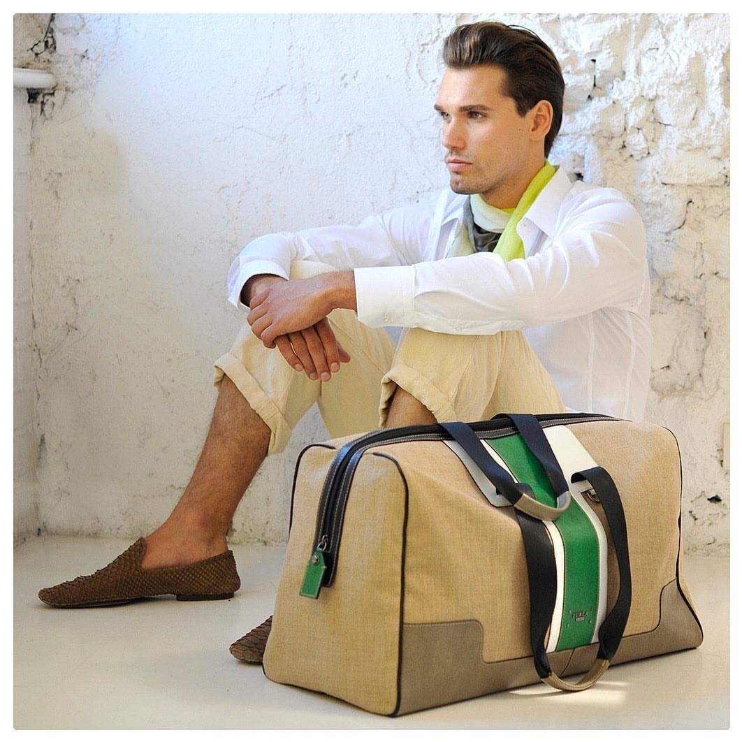 Man in white shirt and beige shorts sitting next to a large tan canvas duffel bag.