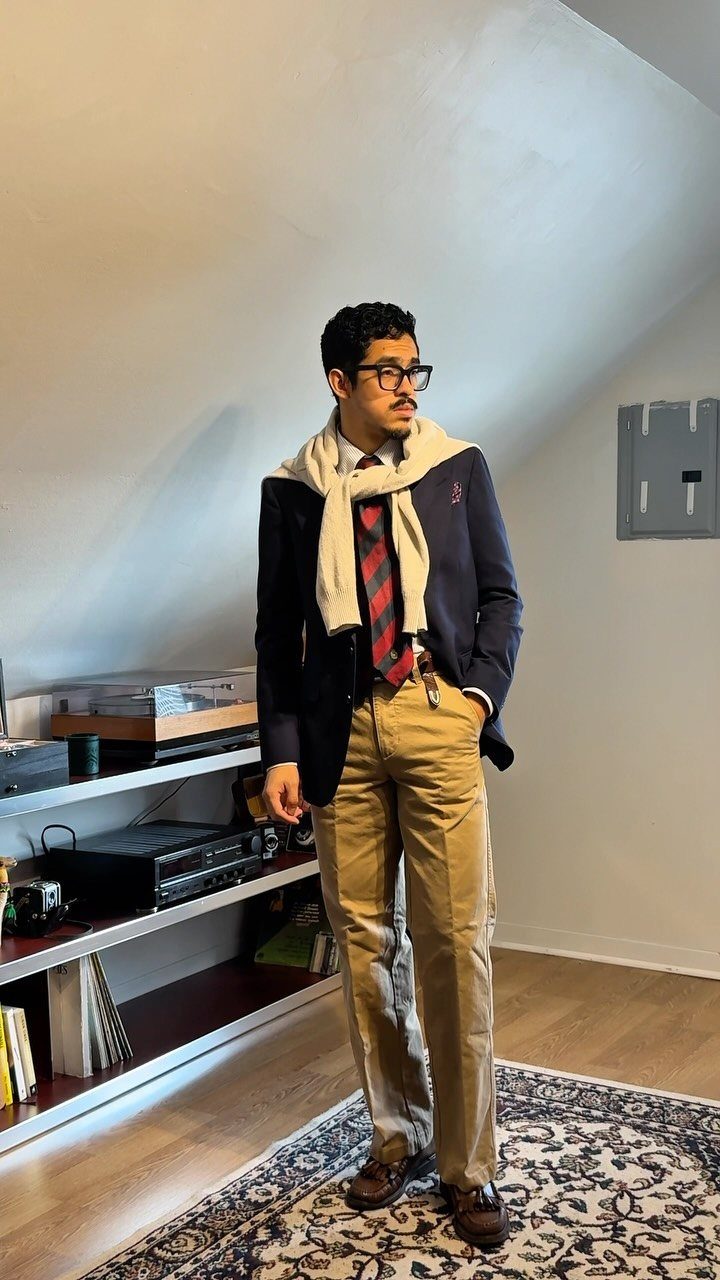 A man posing in a navy blazer, striped tie, tan trousers, and a sweater draped over his shoulders.