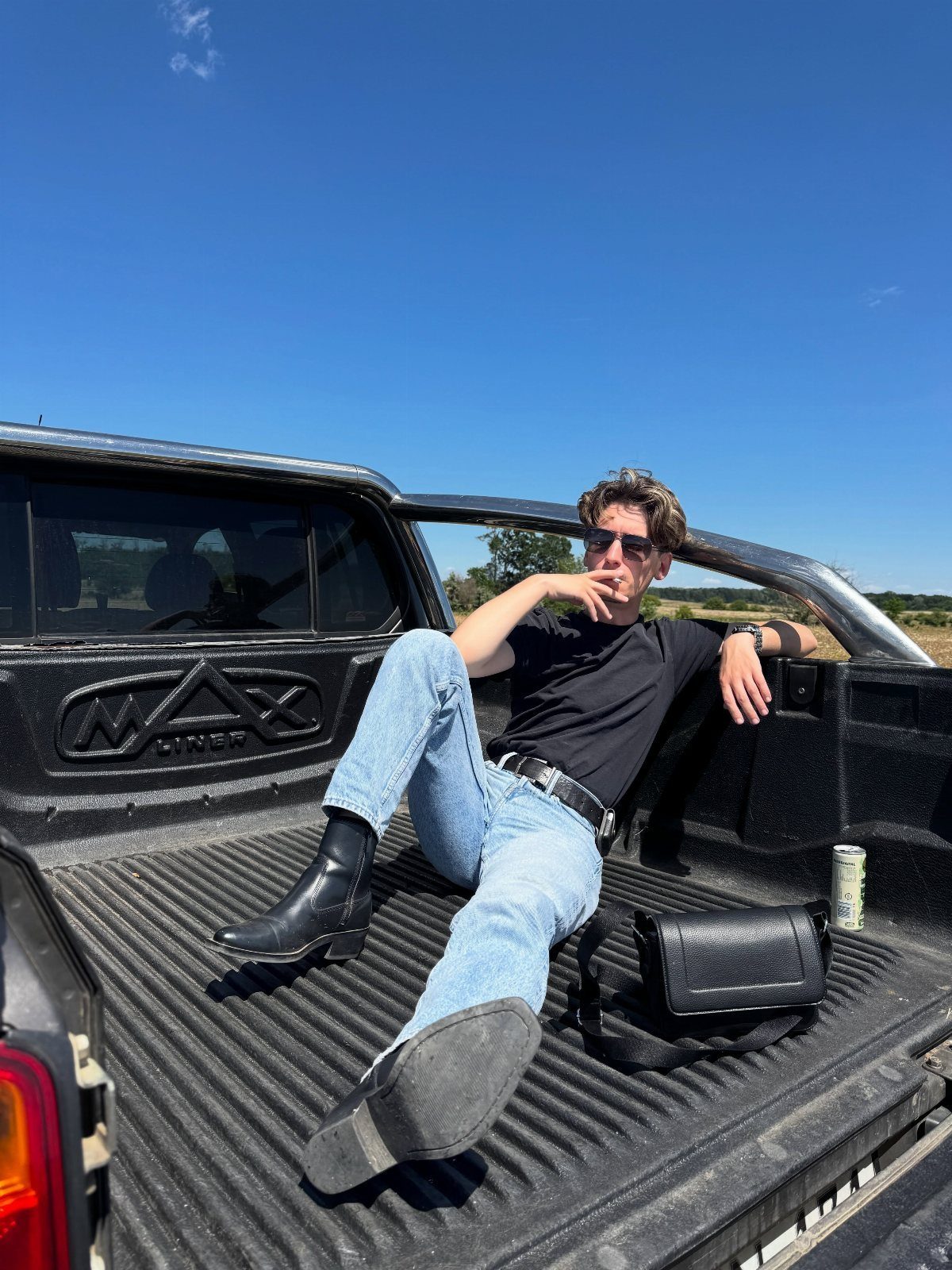A man in a black t-shirt and blue jeans sitting in a black pickup truck bed.