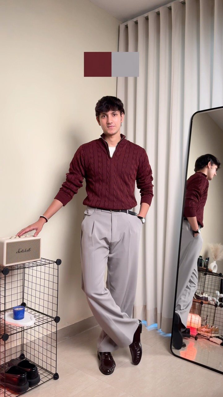 Man wearing a maroon sweater and grey trousers posing in a room near a mirror.