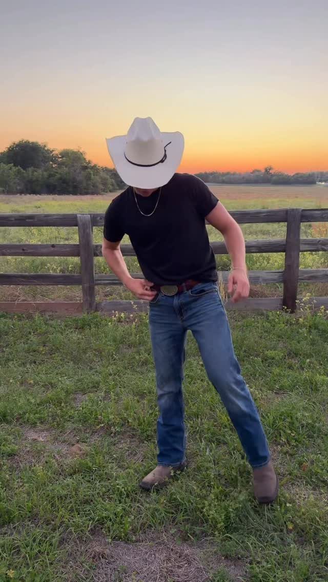 Man in a cowboy hat, black t-shirt, and blue jeans standing in a grassy field at sunset.
