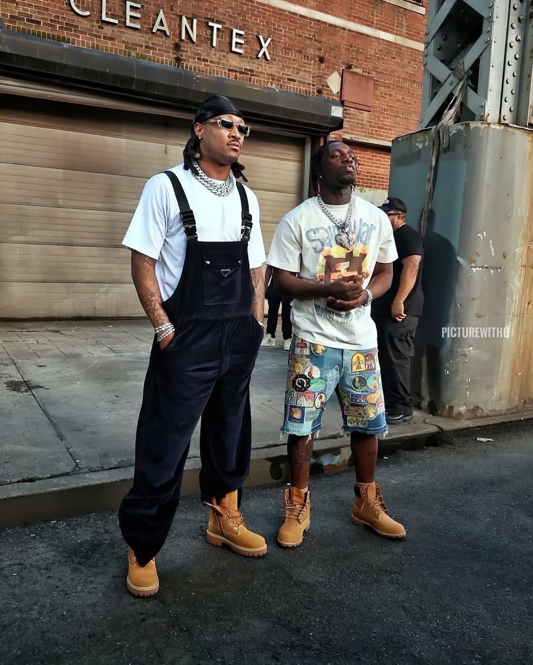Two men posing in streetwear and tan Timberland boots on a city street near a building.