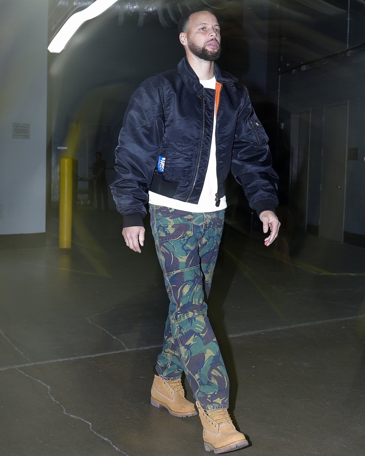 Man walking in a hallway wearing a dark bomber jacket, patterned trousers, and tan Timberland boots.