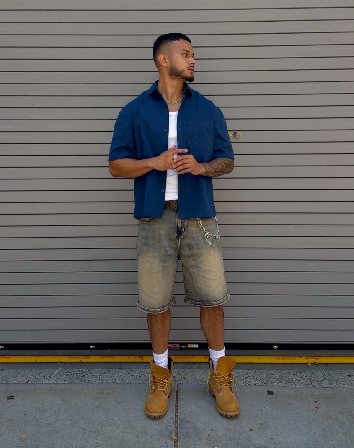 Man wearing a navy button-down shirt, denim shorts, and tan Timberland boots standing outdoors.
