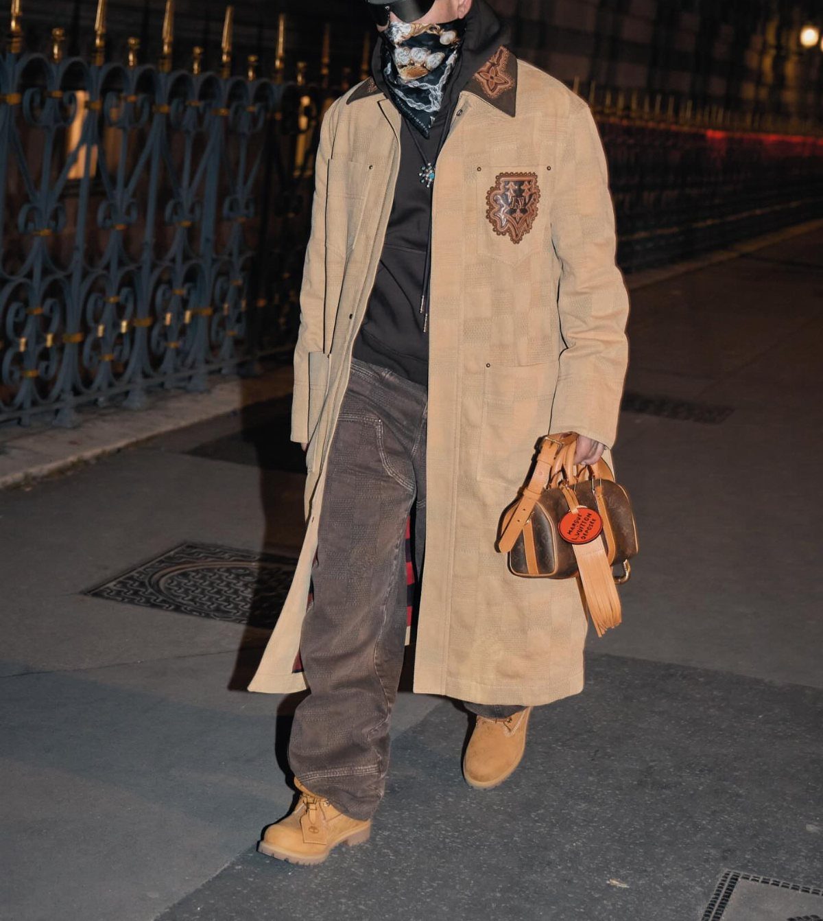 A person wearing a long tan coat, dark trousers, and tan boots walking outdoors at night.
