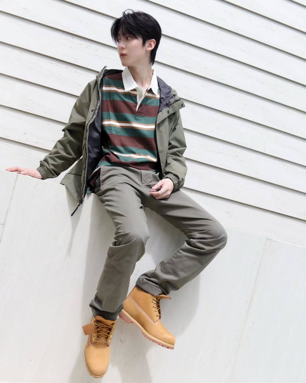 Young man in olive green jacket, striped polo, and tan boots sitting on a ledge.