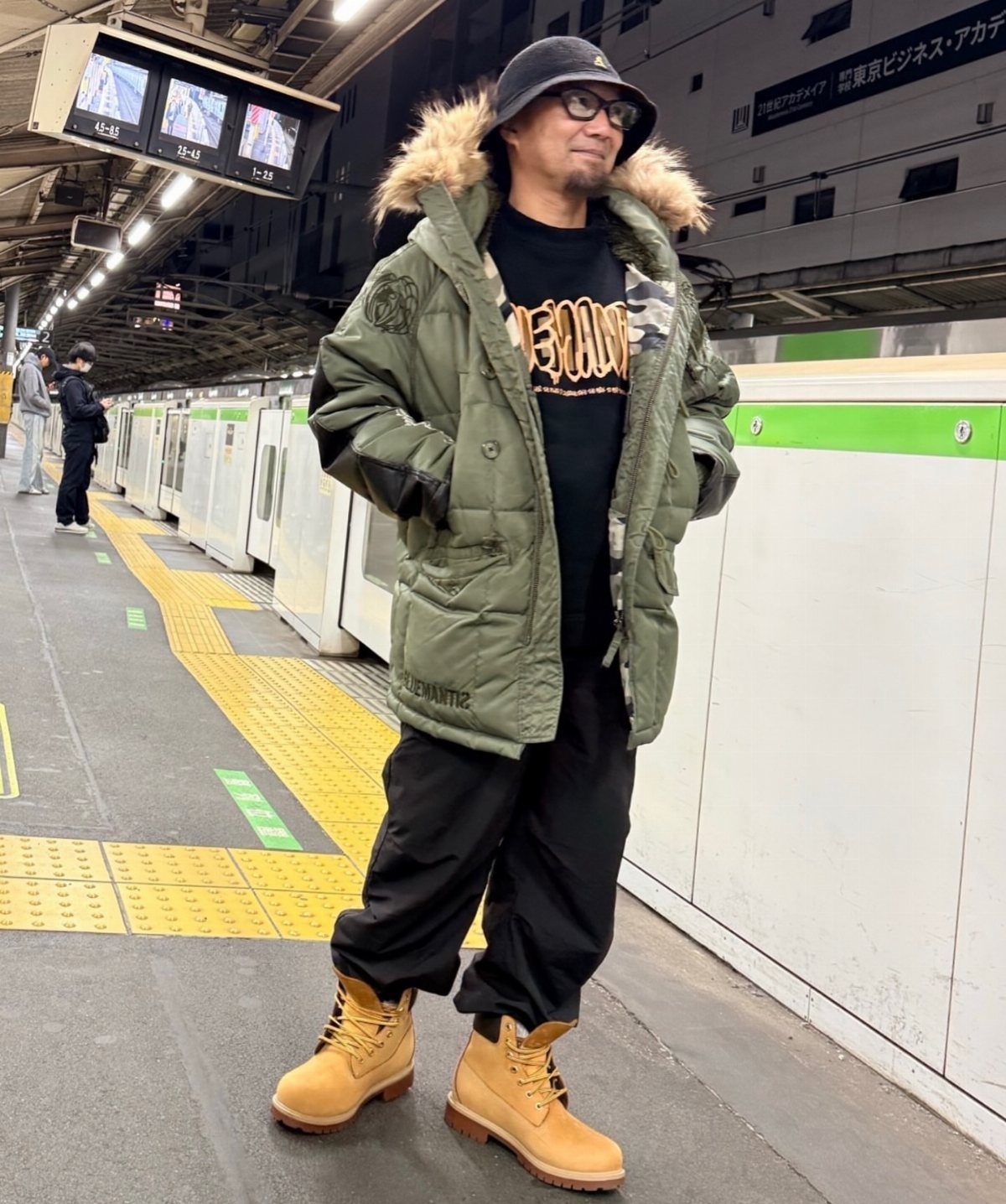 Man in a green puffy parka and tan Timberland boots standing on a train platform.