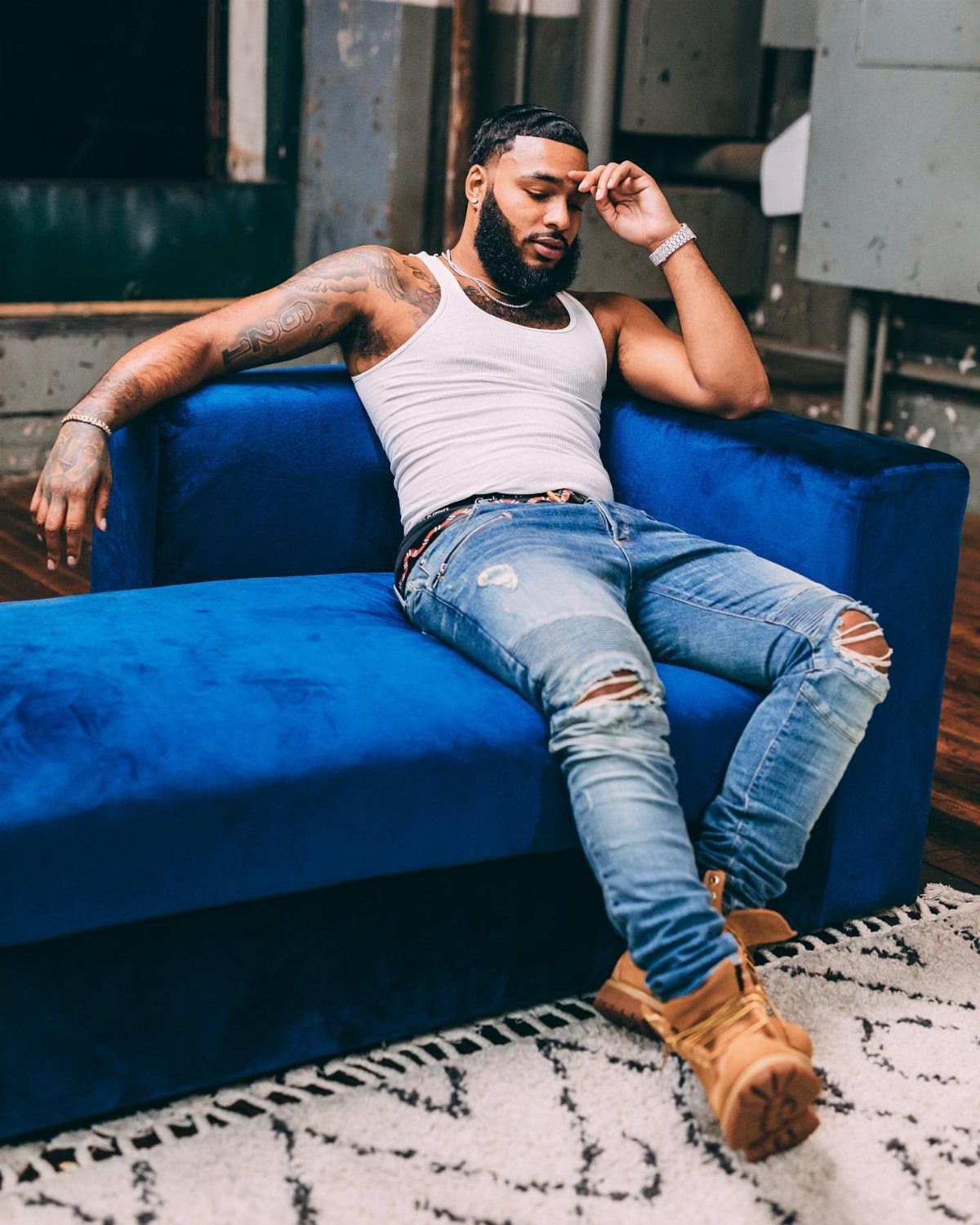 Man in white tank top and ripped jeans sitting on a blue velvet sofa wearing tan boots.