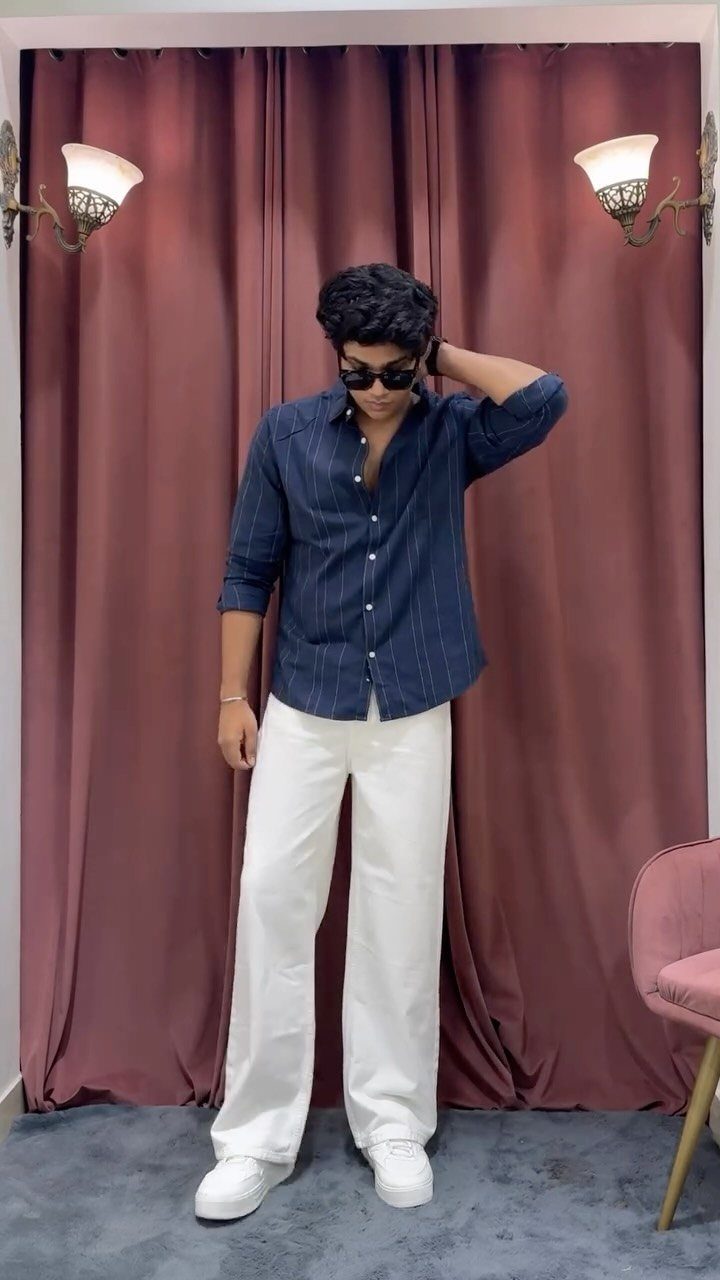 Person wearing a blue striped button-up shirt and wide-leg white jeans standing against mauve curtains.