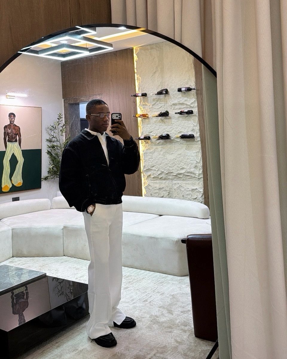 A man posing in a mirror wearing a black jacket and wide-leg white trousers.