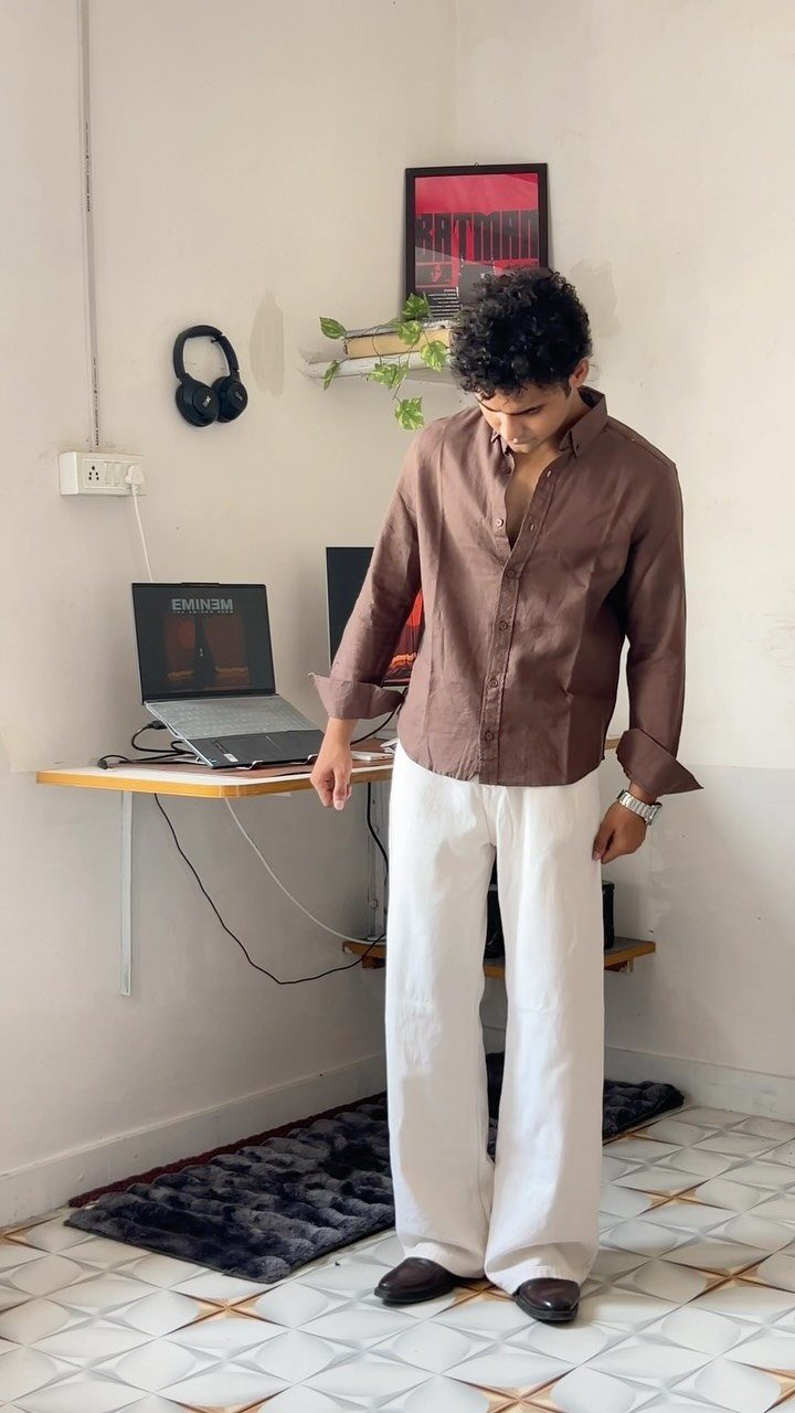 Man wearing a brown button-down shirt and wide-leg white trousers standing in a room.