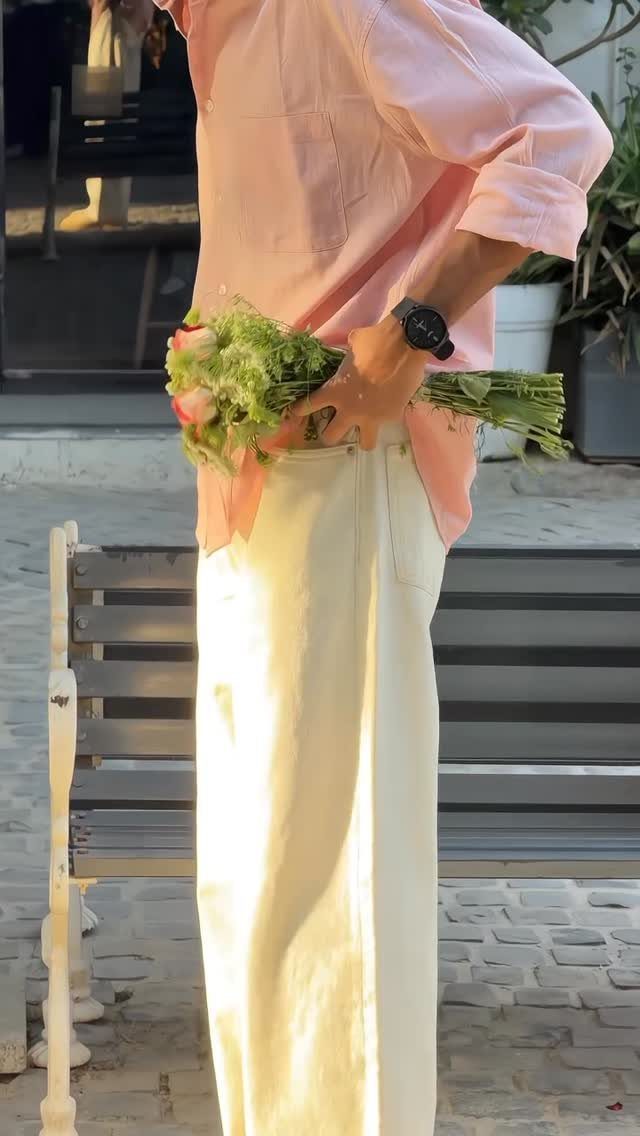 Person in a pink shirt and cream wide-leg pants holding flowers behind their back.