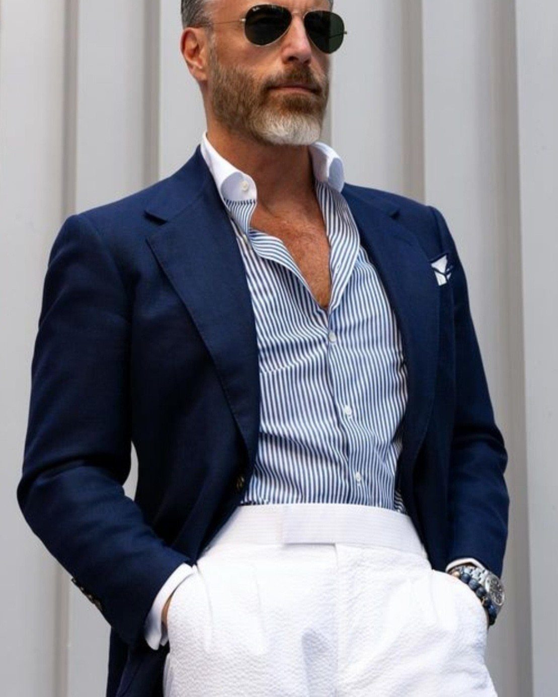 Man in a navy blazer, striped blue and white shirt, and white trousers wearing sunglasses.