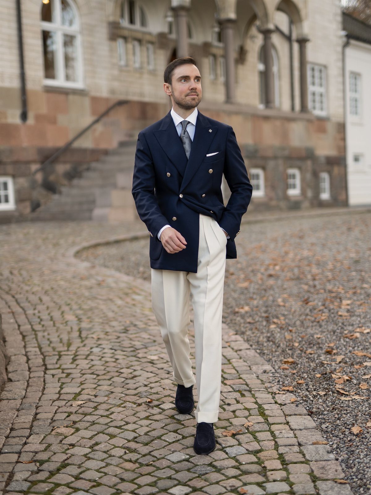 Man walking outdoors wearing a navy blazer, white shirt, tie, and cream trousers.