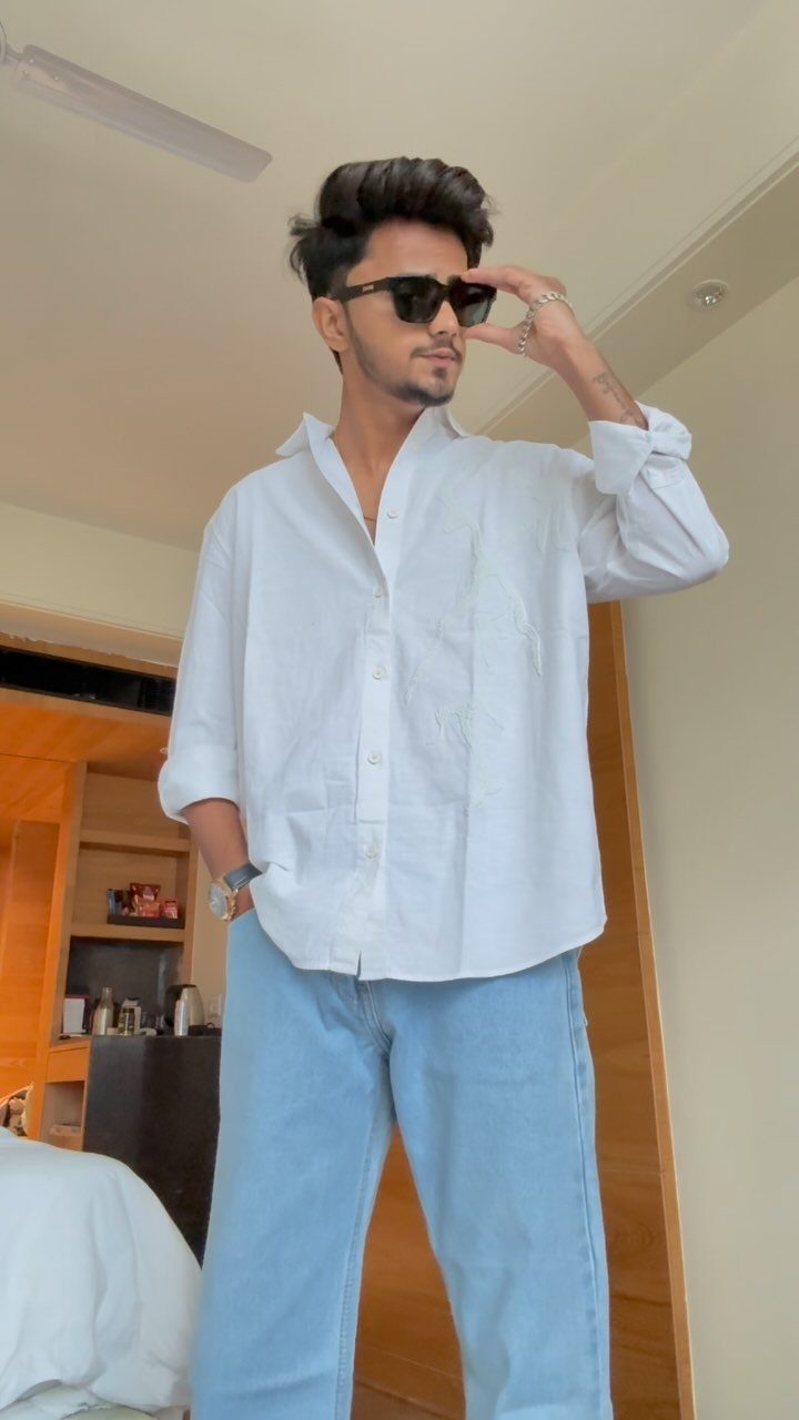 A man wearing a white button-down shirt and light blue jeans while posing indoors.