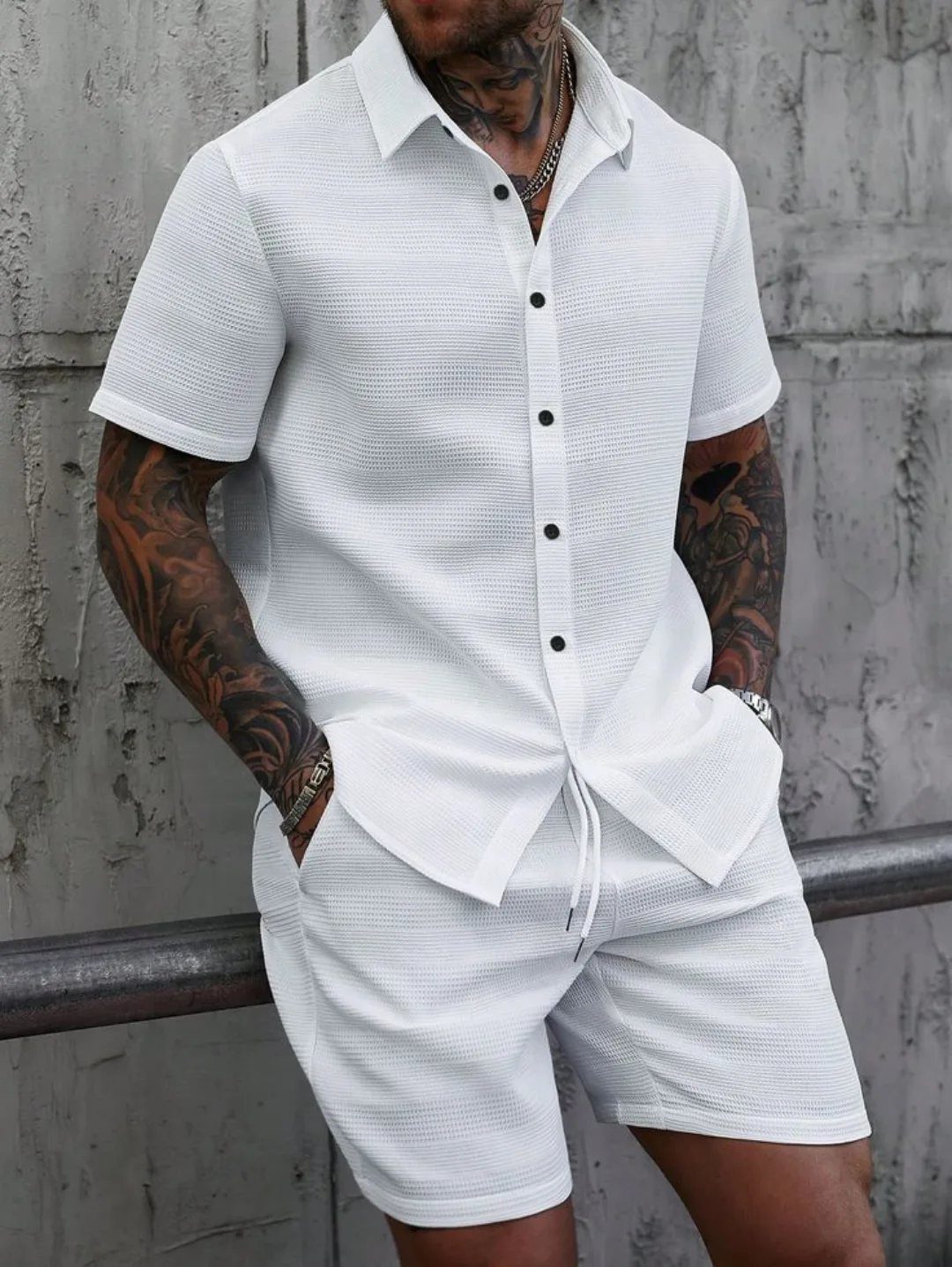 Man wearing a white short-sleeved button-down shirt and matching white shorts against a concrete wall.