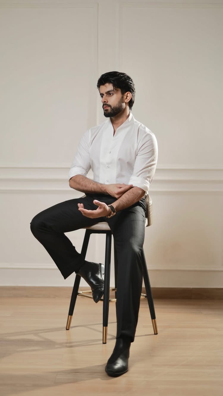 Man in a white shirt tucked into black tailored trousers sitting on a stool.