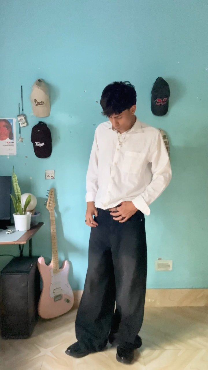 A man wearing a white shirt and black wide-leg trousers standing against a blue wall.