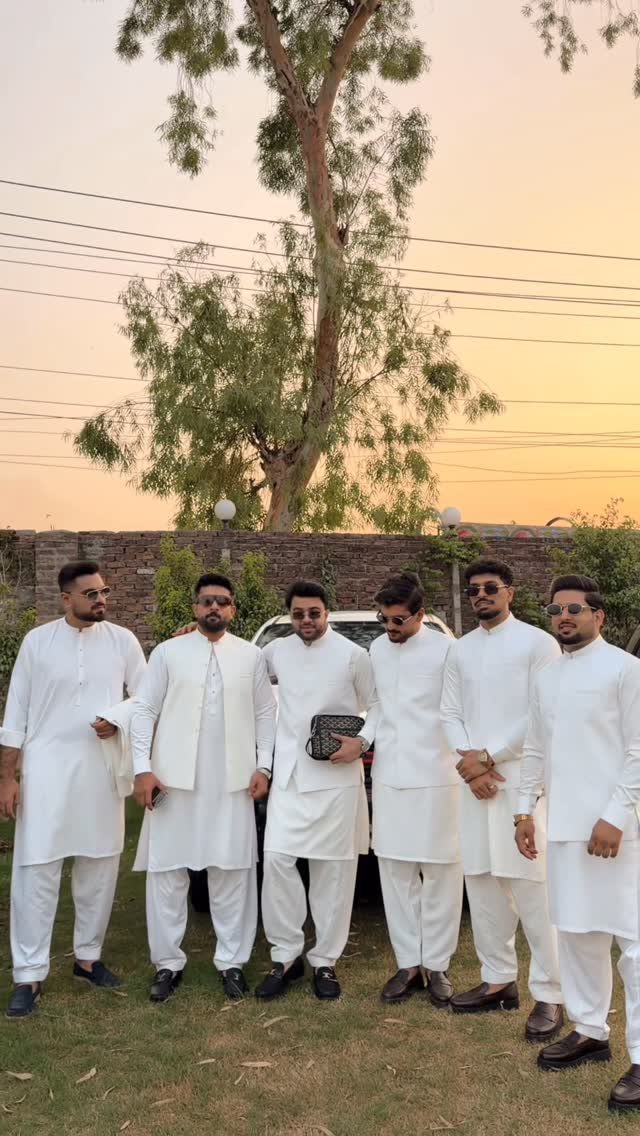Seven men posing together wearing matching white traditional outfits and vests outdoors at sunset.