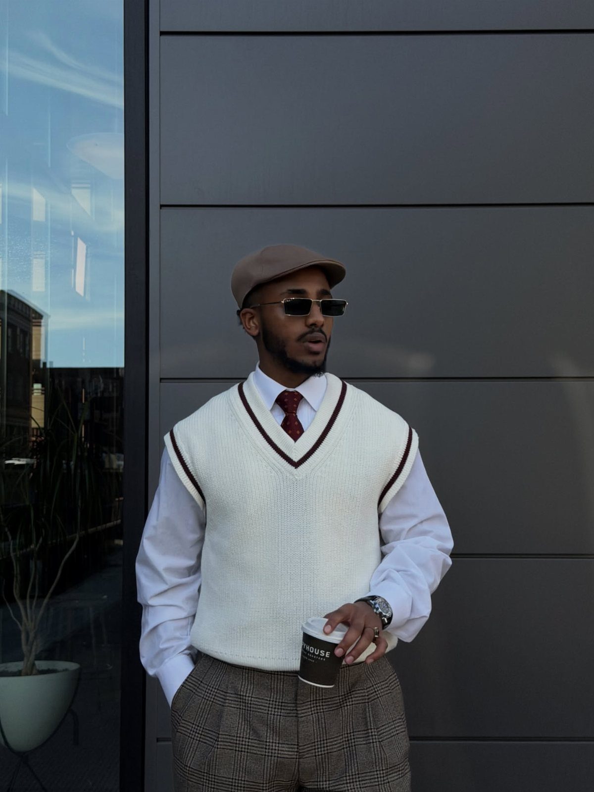 Man wearing a white knitted sweater vest, white shirt, red tie, and tan flat cap.