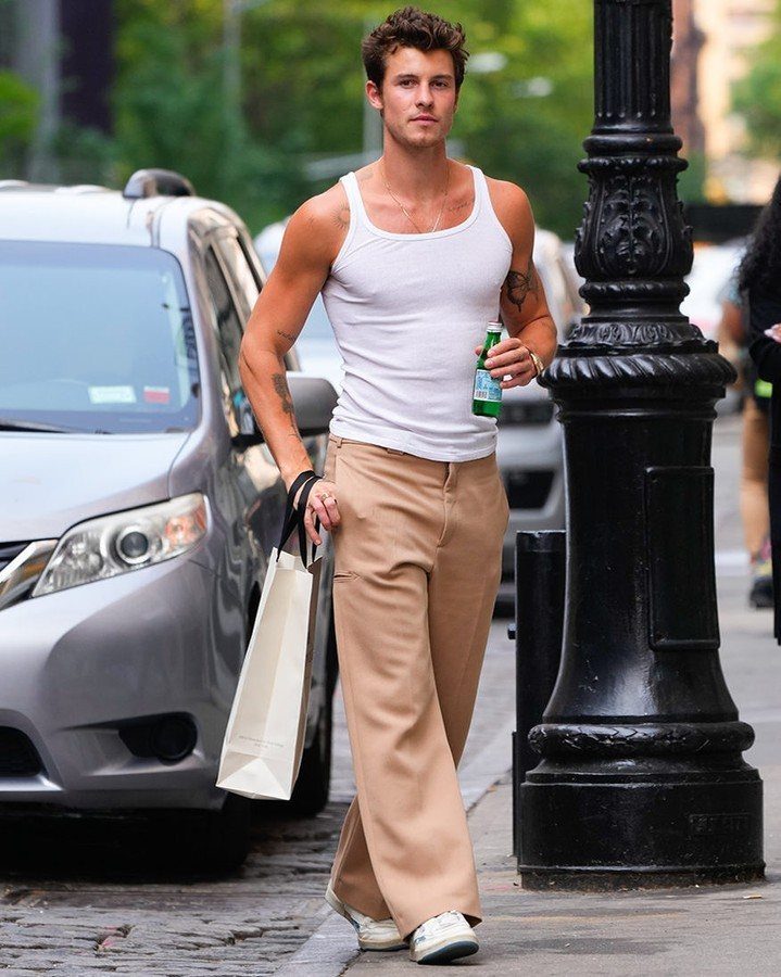 A man walking on a city street wearing a white tank top and tan trousers.