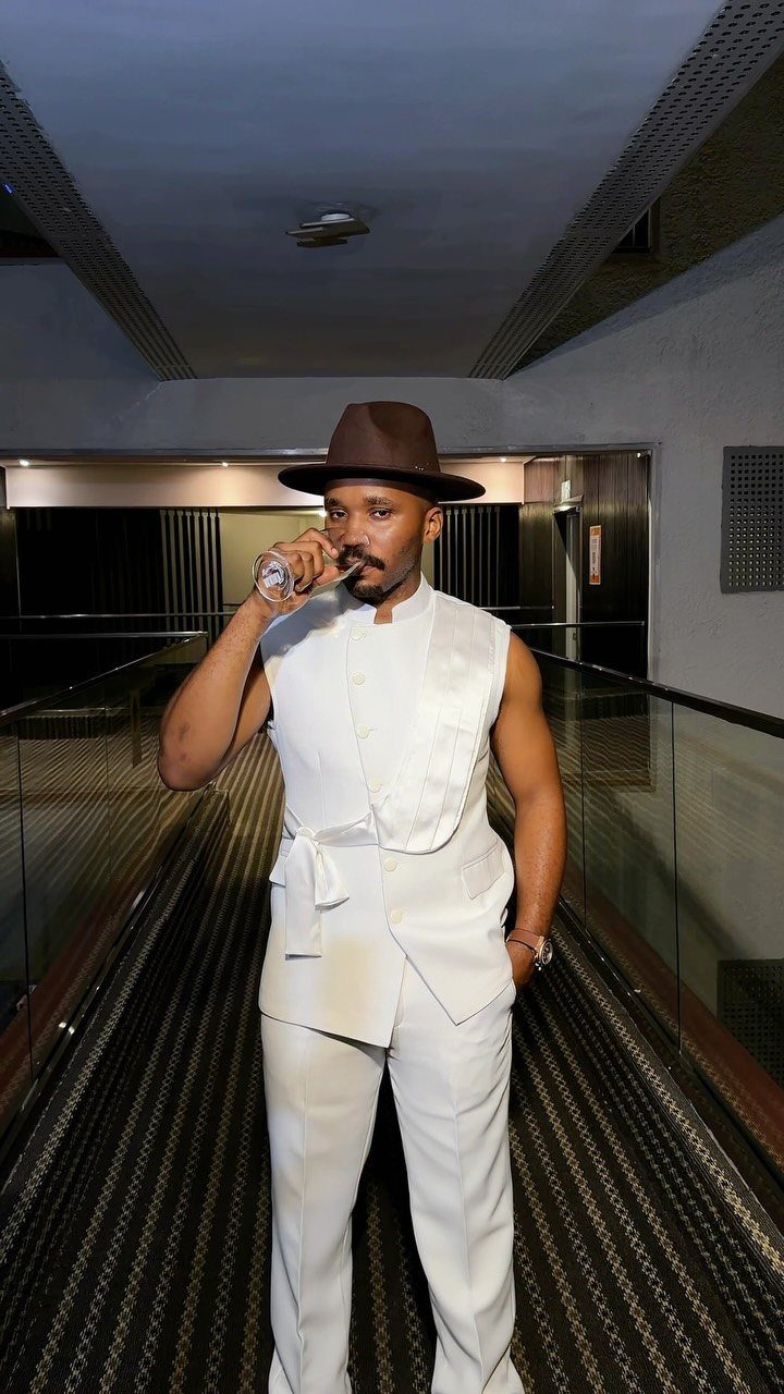 A man in a white vest and trousers posing in a hallway wearing a brown hat.